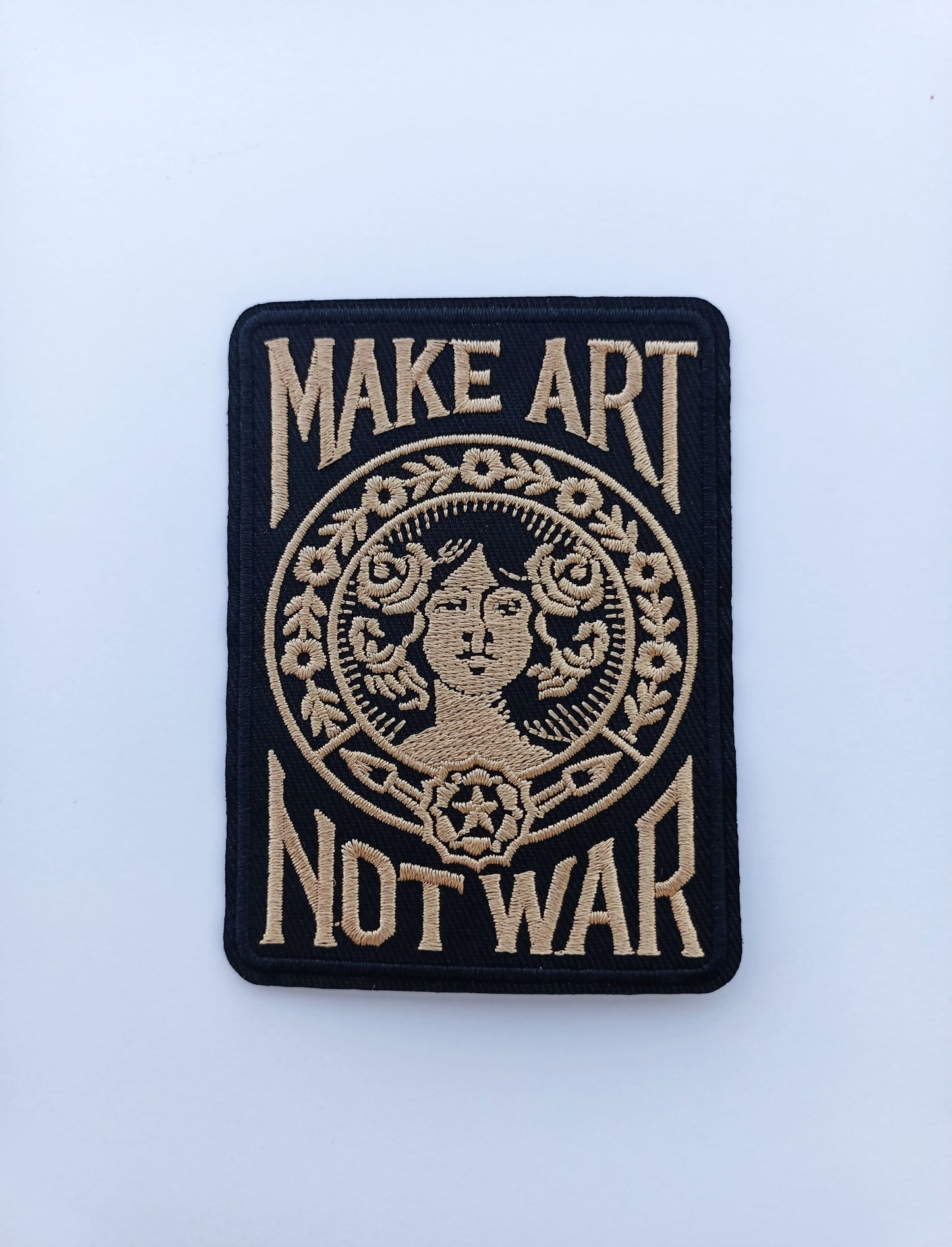 Parche de tela con bordado en contraste que muestra el lema Make Art Not War y figura central femenina.
