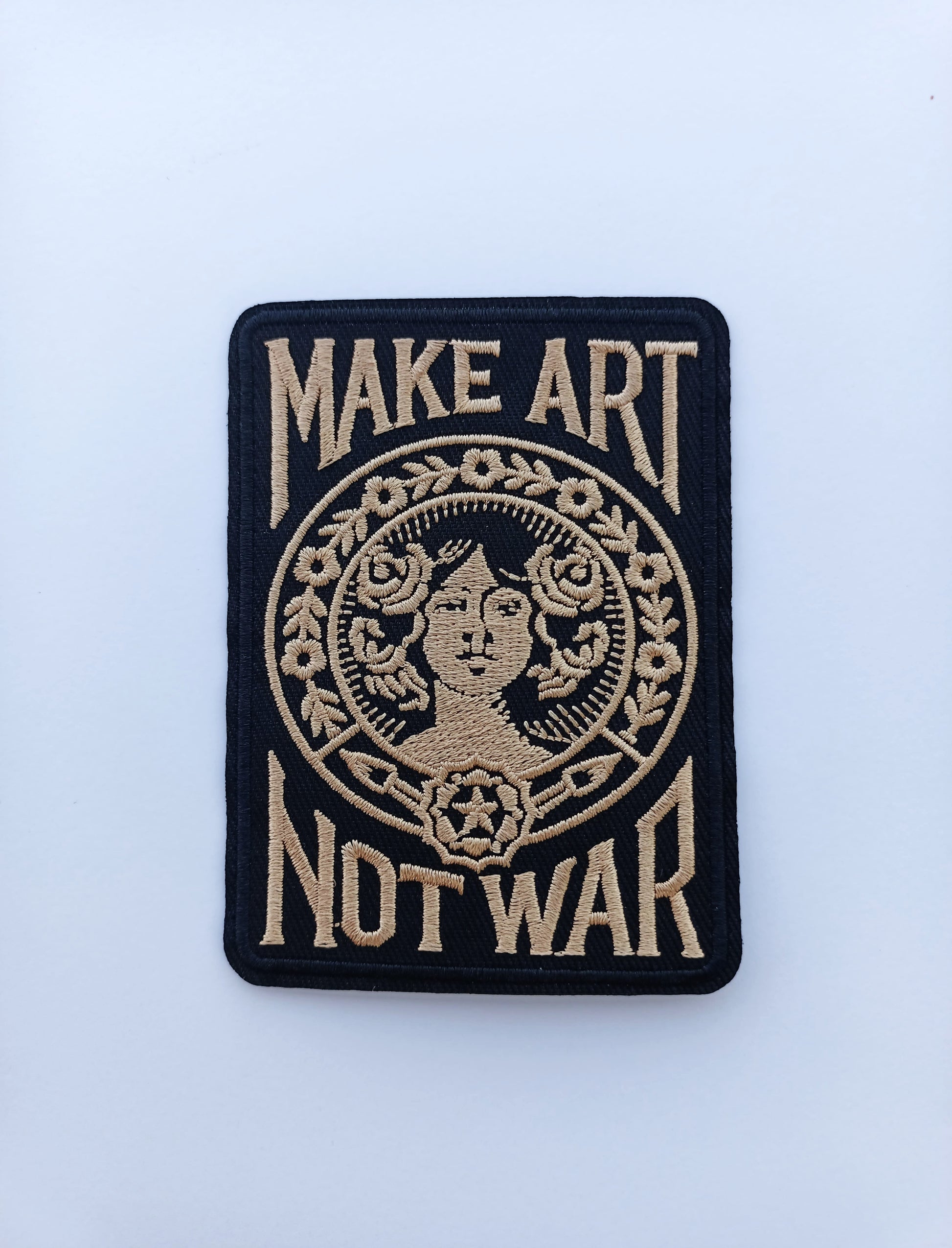 Parche de tela con bordado en contraste que muestra el lema Make Art Not War y figura central femenina.